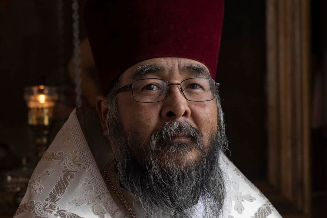 Orthodox Christian priest in a red hat and white vestments with a long gray beard, standing in a church with lit candles and religious icons in the background.