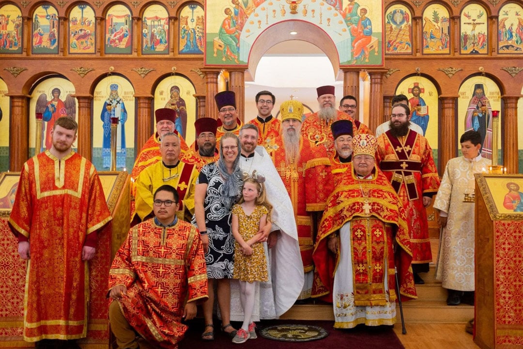 Group of Orthodox Christian clergy in colorful vestments, standing with a family in front of an iconostasis with religious icons in a church.