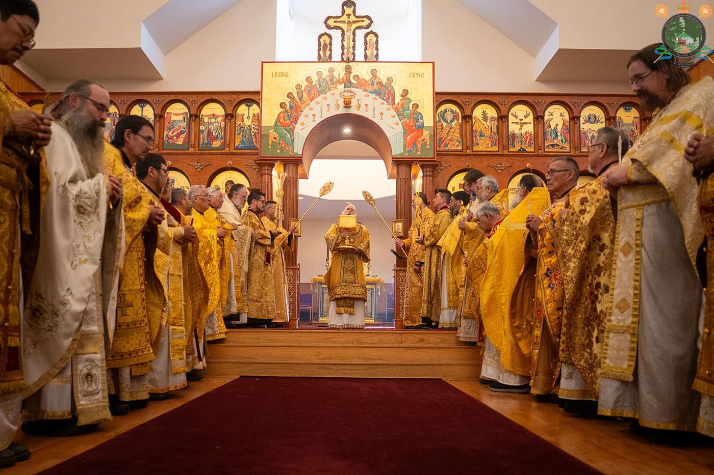 A group of clergy in ornate golden vestments during a religious ceremony inside a church, with a central figure holding large candles and an iconostasis featuring religious icons in the background.