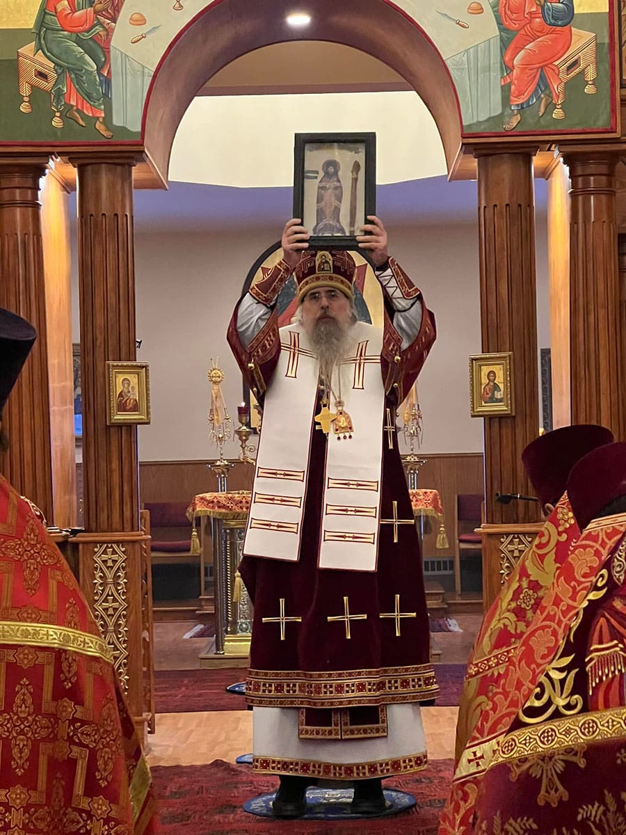 An Orthodox clergy member in red and white vestments holding a framed icon during a service, standing in front of a wooden iconostasis with religious icons.