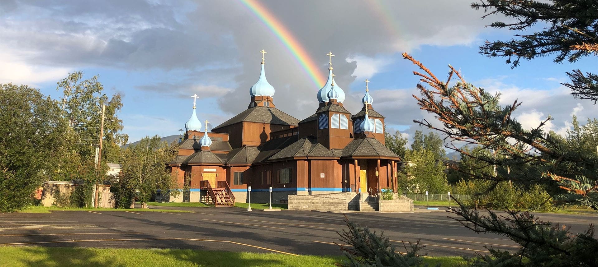 A wooden Orthodox church with blue onion domes under a partly cloudy sky, with a rainbow arching above and green trees in the background.