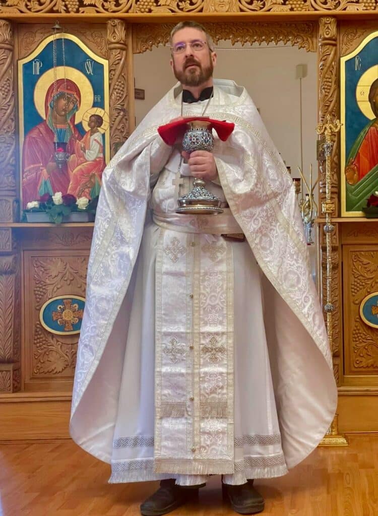 An Orthodox clergy member in white vestments with gold embroidery, holding a chalice covered with a red cloth, standing in front of an iconostasis with icons of the Virgin Mary and Christ.