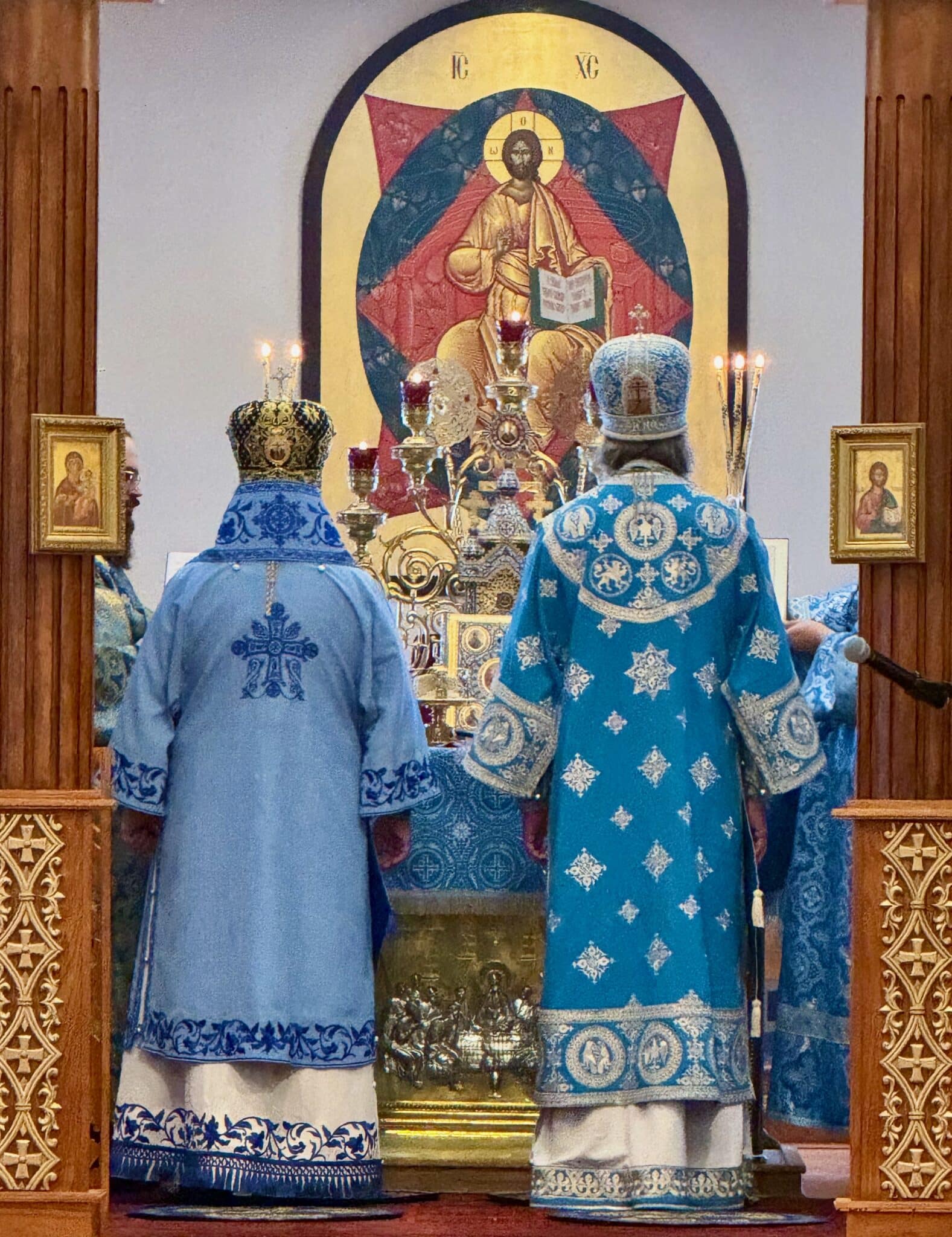 Three Orthodox bishops in ornate blue vestments with gold embroidery perform a service in a church, standing before an icon of Christ and holding liturgical items.