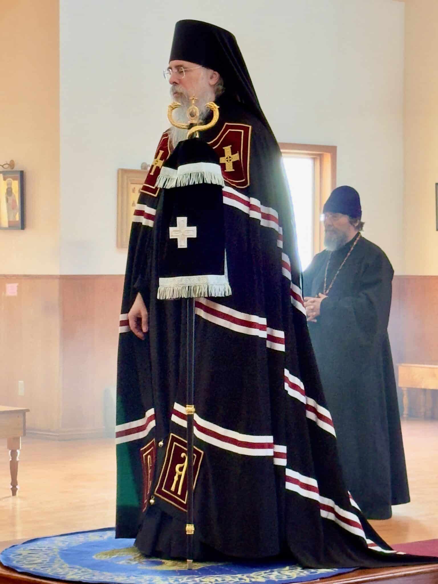 An Orthodox bishop in ornate black and red vestments with gold embroidery stands holding a pastoral staff in a church, with another clergy member in black robes in the background.