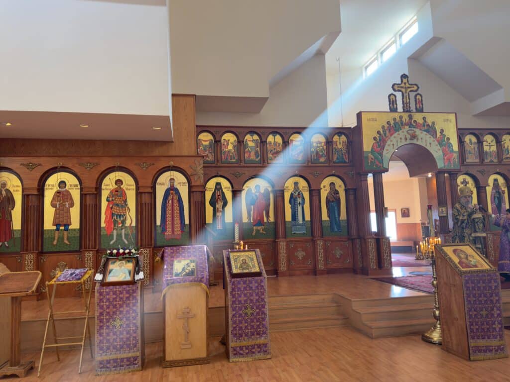 Interior of an Orthodox church with a wooden iconostasis displaying multiple icons of saints, lit candles, and religious artifacts, illuminated by a beam of sunlight from a window.