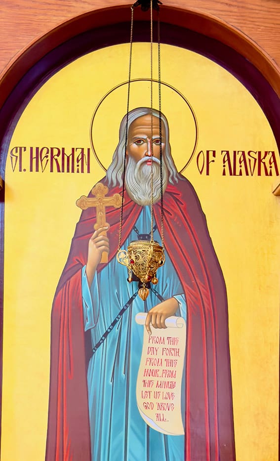 An Orthodox icon of St. Herman of Alaska, depicted with a white beard in blue and red robes, holding a censer and a scroll with a prayer, surrounded by a golden halo.