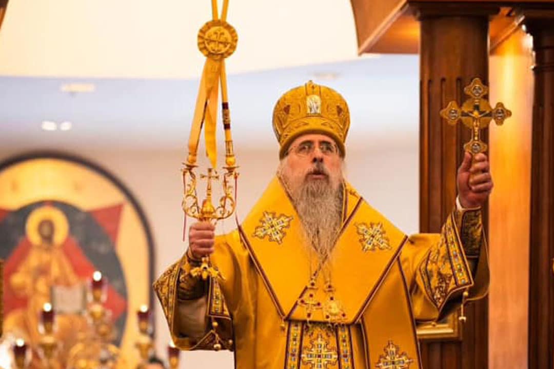 Clergy member in golden liturgical vestments holding a cross and a staff, standing in a church with religious icons in the background.